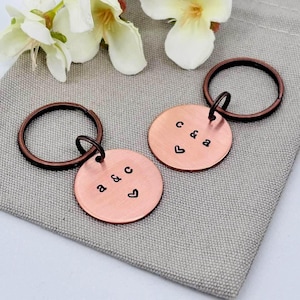 May include: Two circular copper keychains featuring stamped black lettering and a heart symbol. One keychain is stamped with the letters a & c, and the other is stamped with c & a. Each metal disc is attached to a dark copper-toned split ring.