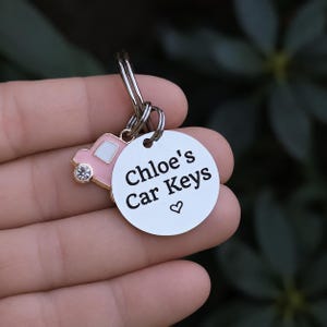 Puede incluir: Un llavero de metal plateado con un disco circular grabado con el texto negro "Chloe's Car Keys" y un peque&ntilde;o s&iacute;mbolo de coraz&oacute;n, acompa&ntilde;ado de un dije de coche en miniatura rosa y blanco con un solo brillante transparente.