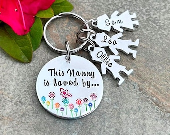 May include: A silver-toned metal keychain featuring a large circular disc engraved with the text This Nanny is loved by and colorful flower and butterfly illustrations. Three smaller silver-toned charms shaped like children are attached to the ring.