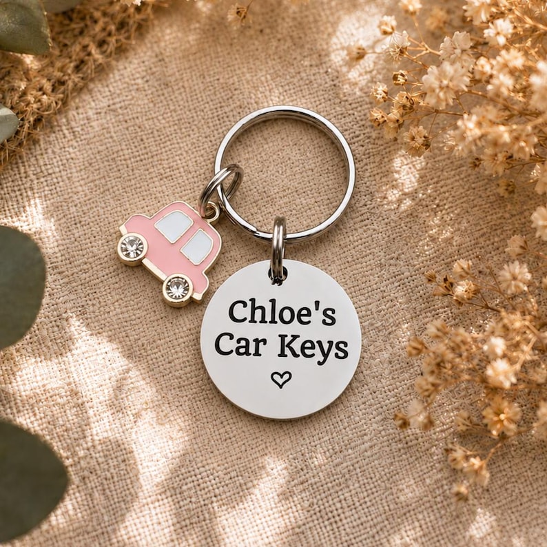 Puede incluir: Un llavero de metal plateado con un disco circular grabado con el texto "Chloe's Car Keys" y un peque&ntilde;o s&iacute;mbolo de coraz&oacute;n, acompa&ntilde;ado de un dije de coche de esmalte rosa con dos cristales transparentes en las ruedas y ventanas blancas.