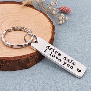 May include: A silver-coloured rectangular metal keyring featuring a split ring and a flat pendant. The pendant is engraved with the black text "drive safe I love you" followed by a small heart symbol, creating a clear and legible message.
