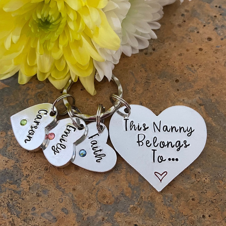 May include: A silver heart-shaped keychain with the text "This Nanny Belongs To..." and a small heart. There are three smaller heart-shaped charms attached to the keychain with the words "Carson", "Faith", and "Emily" and small colorful gemstones.