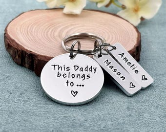May include: Silver keychain with a round charm that says "This Daddy belongs to..." with a heart. Two smaller charms with the names "Mason" and "Amelie" are attached to the keychain.