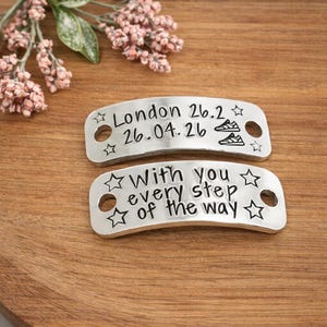 May include: Two curved silver-toned metal shoe tags with holes on each end. The top tag features the text London 26.2, 26.04.26, and two small trainer icons. The bottom tag features the text With you every step of the way and star icons.