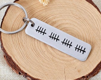 May include: A rectangular silver-toned metal keychain featuring a brushed finish and a black engraved tally mark design consisting of four groups of five lines. The keychain is attached to a circular silver metal split ring for keys.