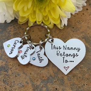 May include: Silver heart-shaped keychain with the text "This Nanny Belongs To..." and three smaller heart-shaped charms with the names "Carson", "Emilee", and "Faith".