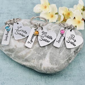 Puede incluir: Tres llaveros de metal plateado, cada uno con un dije en forma de coraz&oacute;n grabado con Little Sister, Middle Sister o Big Sister, junto a una etiqueta rectangular grabada con los nombres Sophie, Lynsey o Jenny y un peque&ntilde;o cristal de color.