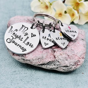 May include: A silver-toned metal keychain featuring a large circular disc engraved with the text My Weight Loss Journey and a heart symbol. Three smaller heart-shaped charms are attached, each stamped with a number and the word stone.