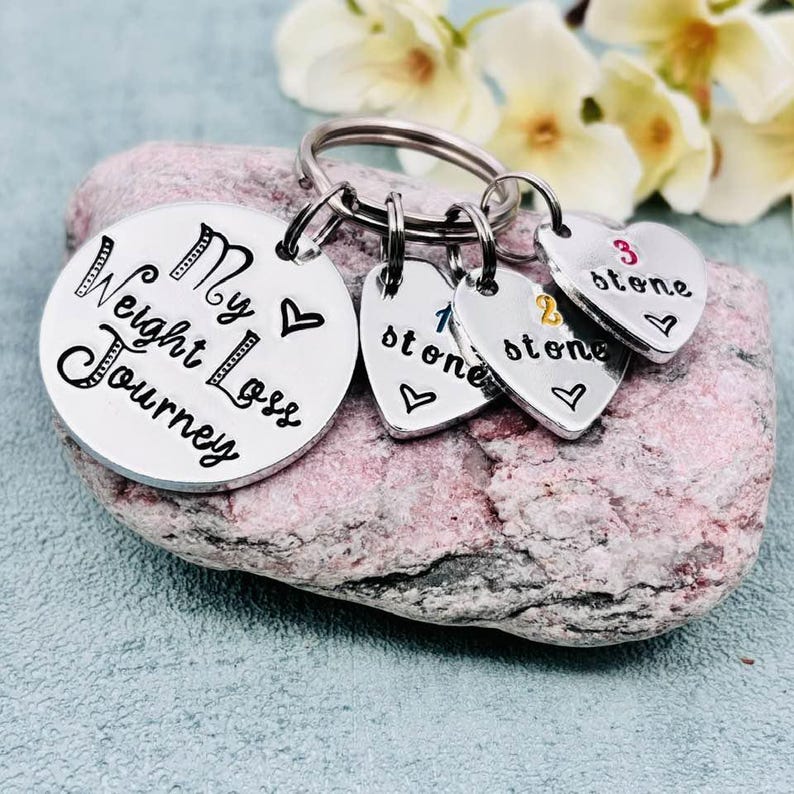 May include: A silver-toned metal keychain featuring a large circular disc engraved with the text My Weight Loss Journey and a heart symbol. Three smaller heart-shaped charms are attached, each stamped with a number and the word stone.
