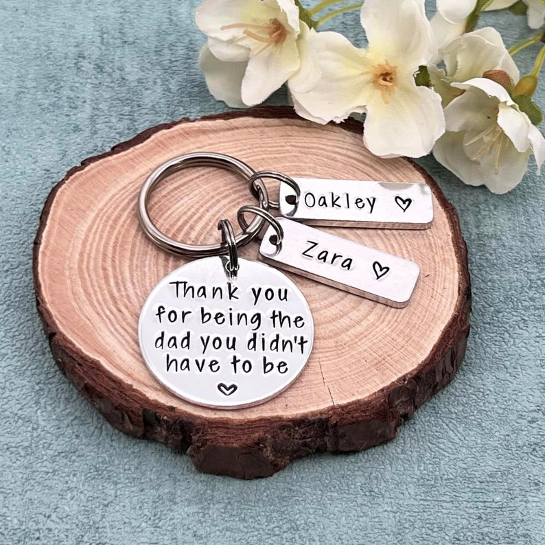 May include: Silver keychain with a round charm that reads "Thank you for being the dad you didn't have to be".  The keychain also has two smaller charms with the names "Oakley" and "Zara" on them.