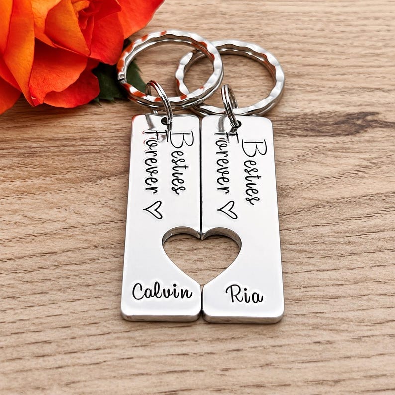 May include: Two silver-toned metal rectangular keychains featuring a split heart cutout design. Each piece is engraved with the text Besties Forever, a small heart symbol, and the names Calvin and Ria respectively, attached to circular rings.