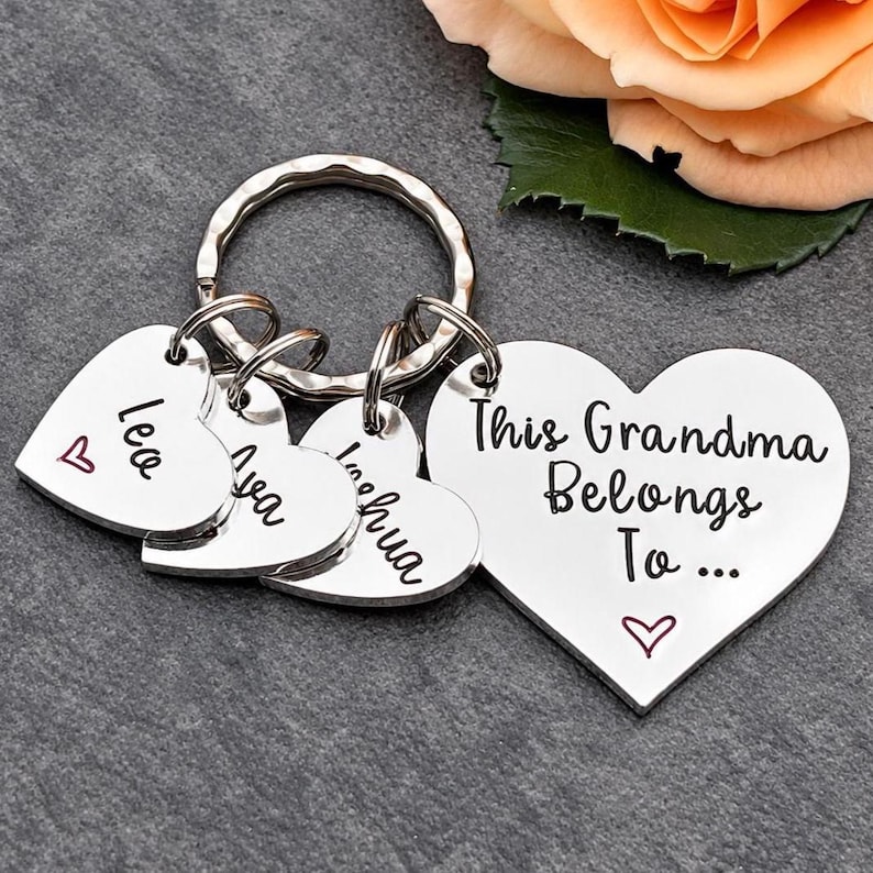 May include: A silver-toned metal keyring featuring a large heart-shaped charm engraved with the text This Grandma Belongs To... and three smaller heart-shaped charms engraved with the names Lea, Ava, and Joshua, each accented with a small red heart.