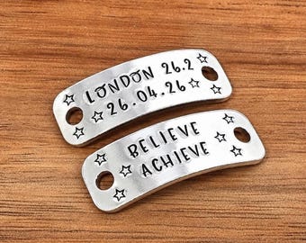May include: Two curved silver-toned metal tags with rounded corners and two circular holes on each end. The top tag is stamped with "LONDON 26.2" and "26.04.26" with star accents. The bottom tag is stamped with "BELIEVE" and "ACHIEVE" with star accents.