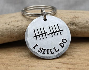 May include: A round silver-toned metal keychain with a hammered edge. The surface features two sets of tally marks engraved in black, with the text I STILL DO stamped in black capital letters below the tally marks and a metal split ring attached.