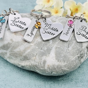 Puede incluir: Tres llaveros de metal plateado, cada uno con una etiqueta rectangular grabada con un nombre y una etiqueta en forma de coraz&oacute;n grabada con Little Sister, Middle Sister o Big Sister. Cada llavero incluye un peque&ntilde;o cristal redondo de color.