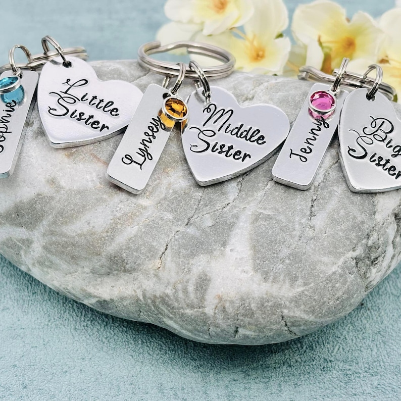 Puede incluir: Tres llaveros de metal plateado, cada uno con una etiqueta rectangular grabada con un nombre y una etiqueta en forma de coraz&oacute;n grabada con Little Sister, Middle Sister o Big Sister. Cada llavero incluye un peque&ntilde;o cristal redondo de color.