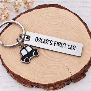 May include: A silver-toned metal keyring featuring a rectangular tag engraved with the black text OSCAR'S FIRST CAR and a small black and white car-shaped charm attached to a circular split ring, all made of durable metal components.