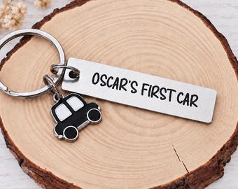 May include: A silver-toned metal keychain featuring a rectangular tag engraved with the black text OSCAR'S FIRST CAR and a small black and white car-shaped charm attached to a circular split ring, all made of durable metal components.