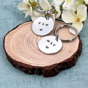 May include: Two silver keychains with stamped initials and a heart symbol. One keychain reads "c & m" and the other reads "m & c". The keychains are on a wooden slice, with white flowers in the background.