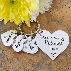 May include: Silver heart-shaped keychain with the text "This Nanny Belongs To..." and a small red heart. Three smaller heart-shaped charms with the names "Carson", "Emily", and "Faith" are attached to the keychain.