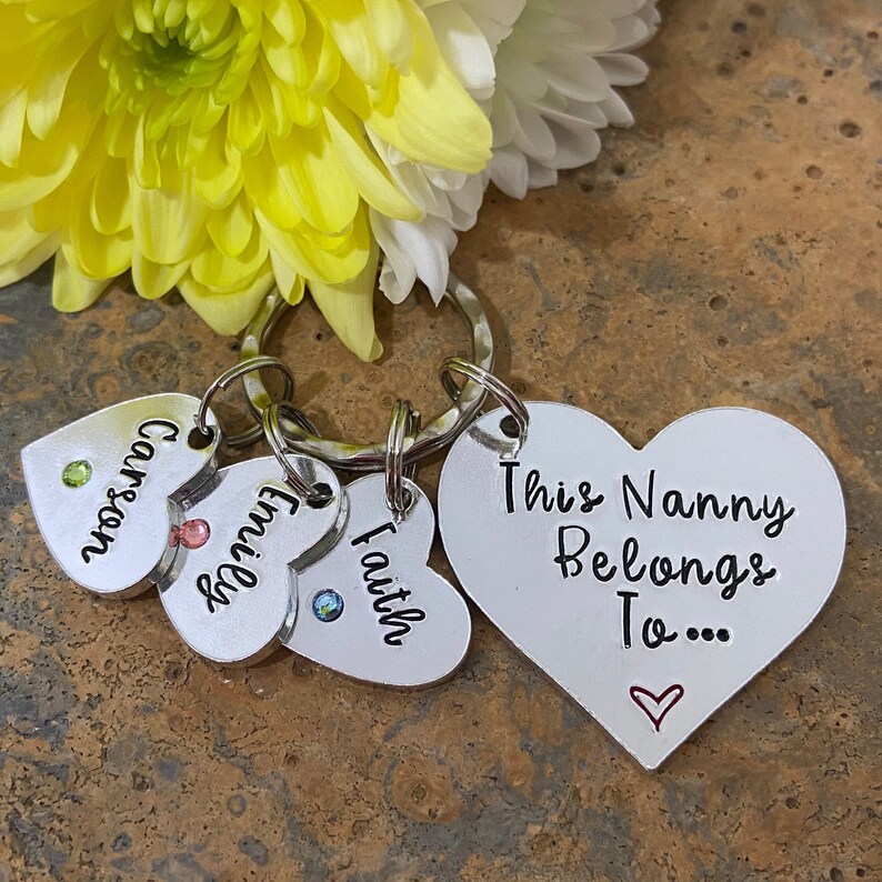 May include: Silver heart-shaped keychain with the text "This Nanny Belongs To..." and a small red heart. Three smaller heart-shaped charms with the names "Carson", "Emilie", and "Faith" are attached to the keychain.