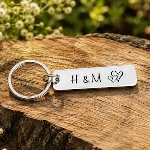 May include: A rectangular silver-toned metal keyring featuring a split ring and a flat tag engraved with the letters H & M followed by two small heart outlines. The metal surface has a smooth, polished finish with clear black lettering.