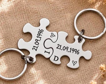 May include: Two silver-toned metal puzzle piece keychains connected together. Each piece is engraved with the date 21.09.1996, the letters I and D, and small heart symbols. Each puzzle piece features a circular metal ring for attaching keys.