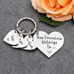 Pode incluir: Um porta-chaves de metal prateado com um pingente grande em forma de cora&ccedil;&atilde;o gravado com o texto This Grandma Belongs To... e tr&ecirc;s pingentes menores em forma de cora&ccedil;&atilde;o gravados com os nomes Leo, Ava e Joshua, cada um com um pequeno cora&ccedil;&atilde;o vermelho.