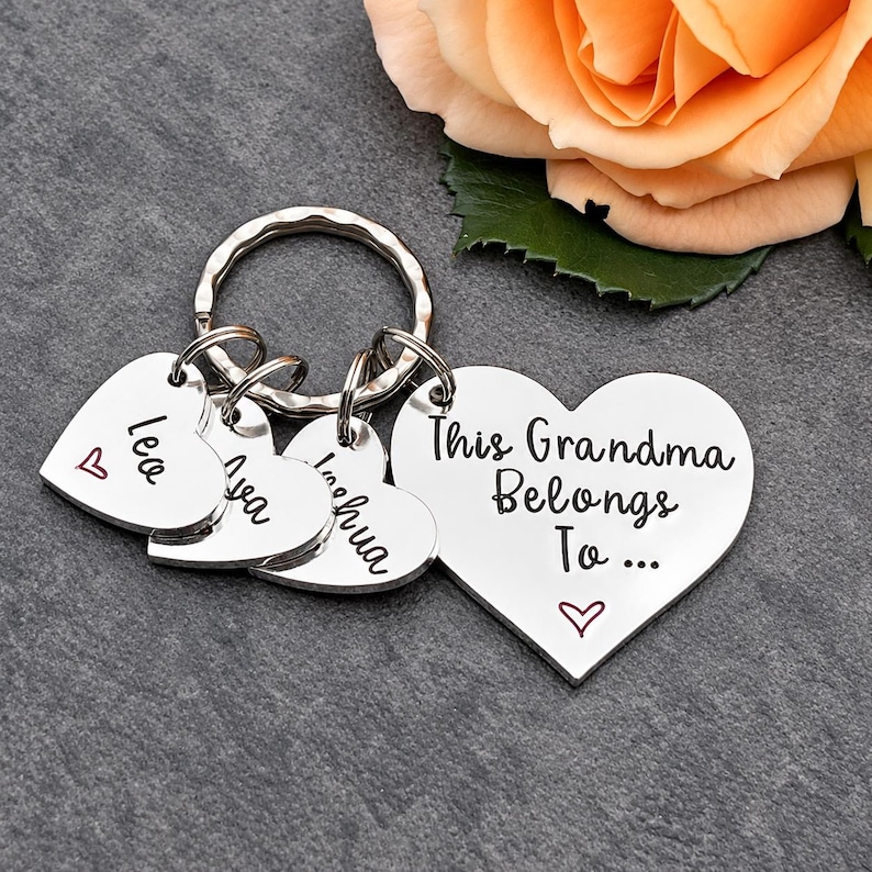Pode incluir: Um porta-chaves de metal prateado com um pingente grande em forma de cora&ccedil;&atilde;o gravado com o texto This Grandma Belongs To... e tr&ecirc;s pingentes menores em forma de cora&ccedil;&atilde;o gravados com os nomes Leo, Ava e Joshua, cada um com um pequeno cora&ccedil;&atilde;o vermelho.