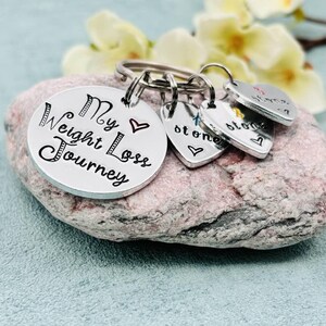 May include: A silver-toned metal keychain featuring a large circular disc engraved with the text My Weight Loss Journey and a small red heart. Three smaller heart-shaped charms are attached, each engraved with the word stone and a colored heart.