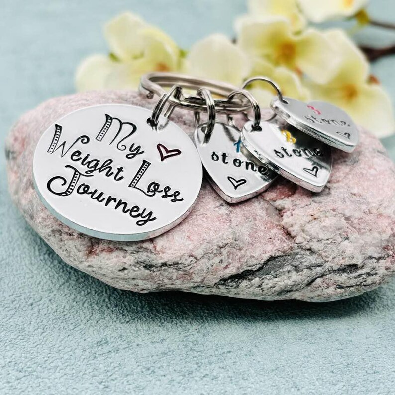 May include: A silver-toned metal keychain featuring a large circular disc engraved with the text My Weight Loss Journey and a small red heart. Three smaller heart-shaped charms are attached, each engraved with the word stone and a colored heart.