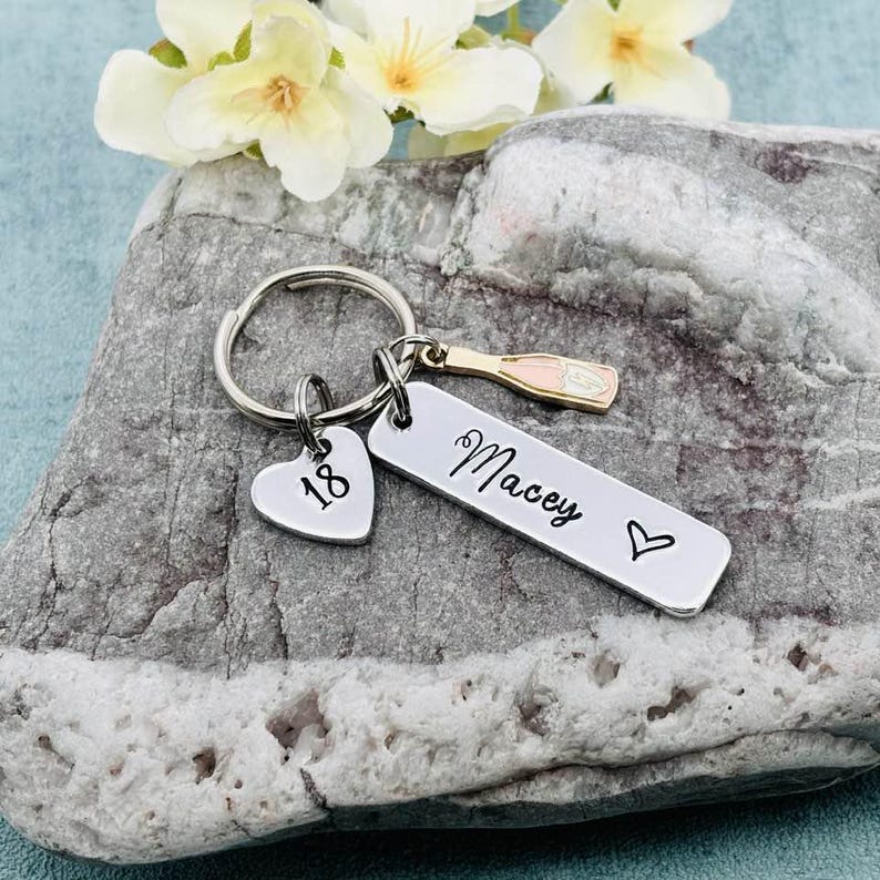 May include: Silver keyring with a heart charm that says "18", a charm of a champagne bottle, and a bar charm that says "Macey" with a heart.