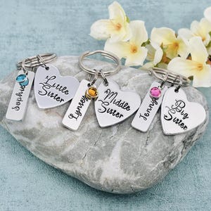 Puede incluir: Tres llaveros de metal plateado, cada uno con una etiqueta rectangular grabada con un nombre y una etiqueta en forma de coraz&oacute;n grabada con Little Sister, Middle Sister o Big Sister. Cada llavero incluye un peque&ntilde;o cristal circular de color.
