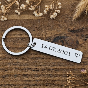 Peut inclure: Un porte-clés en métal argenté composé d'un anneau circulaire attaché à une plaque rectangulaire. La plaque est gravée avec la date 14.07.2001 et un petit symbole de cœur. La surface métallique a une finition lisse et réfléchissante.