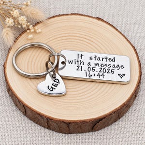 May include: A silver-toned metal keyring featuring a rectangular tag stamped with the text It started with a message 21.05.2025 16:44 and a small heart symbol, accompanied by a heart-shaped charm engraved with the letters G&D on a split ring.