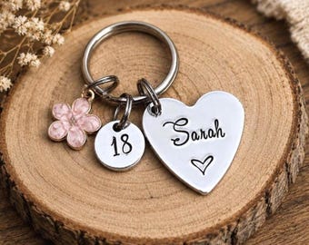 May include: A silver-toned metal keychain featuring three charms: a heart-shaped tag engraved with the name Sarah and a small heart symbol, a circular disc stamped with the number 18, and a five-petaled flower charm with light pink enamel.