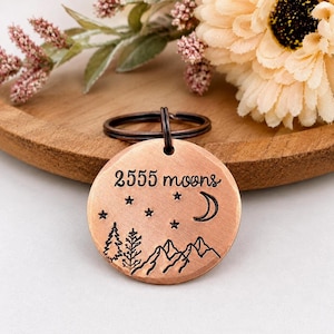 May include: A round copper-coloured metal keyring featuring a stamped design of mountains, two pine trees, stars, and a crescent moon. The text 2555 moons is engraved in black lettering across the top of the circular pendant, which is attached to a dark metal ring.