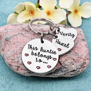 May include: A silver-toned metal keyring featuring a large circular disc engraved with the text This Auntie belongs to... and small red hearts. Two smaller heart-shaped charms are attached, engraved with the names Hazel and Verity in black.