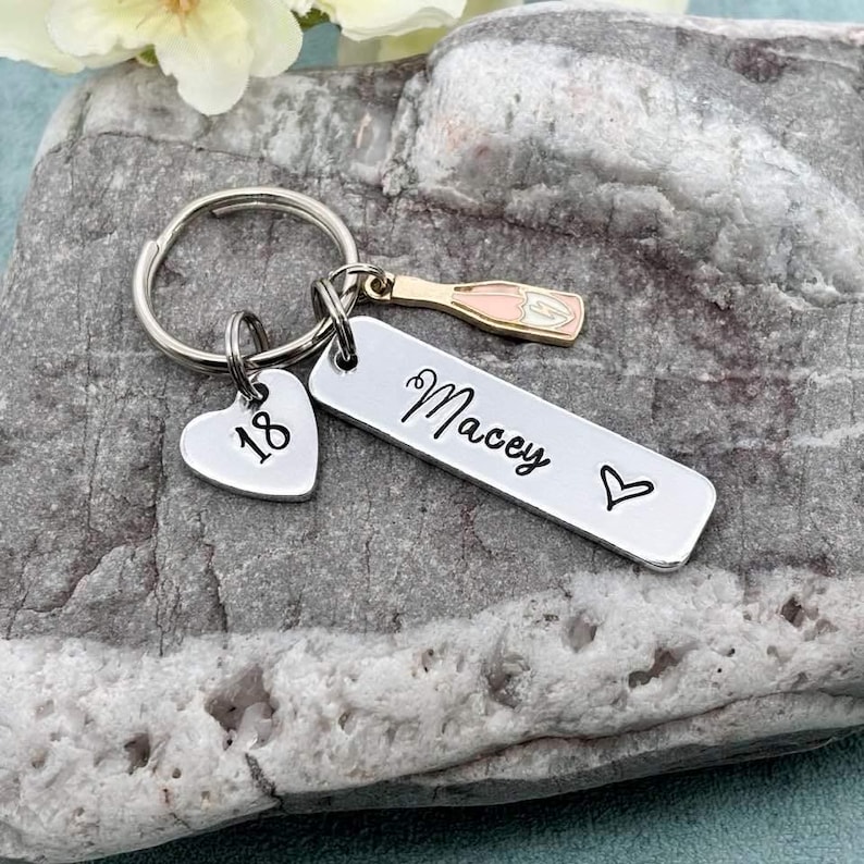 May include: Silver keyring with a heart charm that says "18" and a bar charm that says "Macey" with a small heart. A small charm shaped like a bottle of champagne is attached to the keyring.