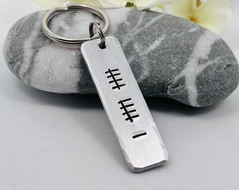 May include: A rectangular, brushed metal keyring with a silver ring. The keyring features a series of vertical lines etched into the metal. The keyring is resting on a grey stone.