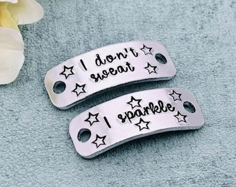 May include: Two silver-toned metal rectangular tags with rounded corners and holes on each end. The top tag features the black engraved text I don't sweat with star icons. The bottom tag features the black engraved text I sparkle with star icons.