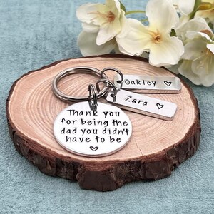 May include: Silver keychain with a round charm that reads "Thank you for being the dad you didn't have to be" and two smaller charms that read "Oakley" and "Zara".