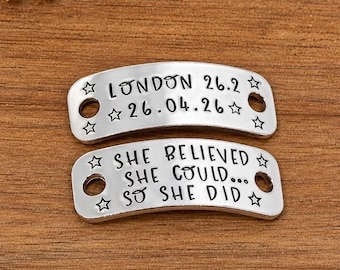 May include: Two curved silver-toned metal tags with holes on each end. The top tag is stamped with "LONDON 26.2" and "26.04.26" surrounded by stars. The bottom tag is stamped with "SHE BELIEVED SHE COULD... SO SHE DID" and star accents.