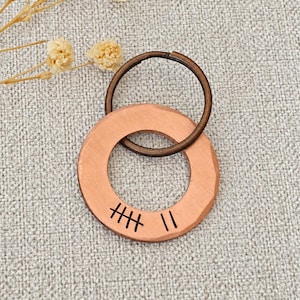 May include: A circular copper pendant with a hammered texture and a dark bronze-toned metal split ring attached. The copper disc features a stamped tally mark design consisting of five lines crossed by a diagonal line and two additional vertical lines.