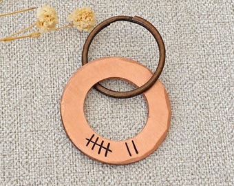 May include: A circular copper pendant with a hammered texture and a dark bronze-toned metal split ring attached. The copper disc features a stamped tally mark design consisting of five lines crossed by a diagonal line and two additional vertical lines.