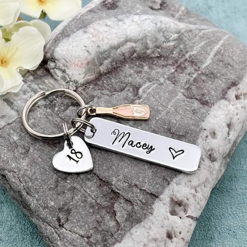 May include: Silver keyring with a heart charm engraved with the number 18, a charm shaped like a bottle of champagne, and a rectangular tag engraved with the name "Macey" and a heart.