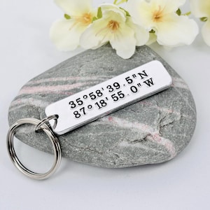May include: A silver rectangular keychain with coordinates 35°58'39.5"N 87°18'55.0"W is displayed on a gray and pink stone, with white flowers in the background. The keychain has a silver key ring attached.