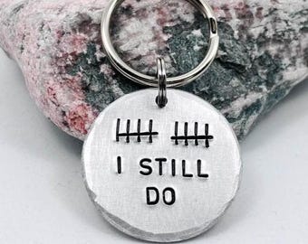 May include: A round silver-toned metal keyring featuring two sets of tally marks stamped at the top and the text I STILL DO stamped in black capital letters below. The keyring includes a circular metal split ring for attachment.