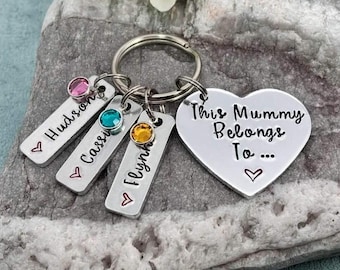 May include: A silver-toned metal keychain featuring a heart-shaped charm engraved with the text This Mummy Belongs To... and three rectangular tags engraved with the names Hudson, Cassi, and Flynn, each accented with a small colored crystal.