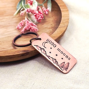 May include: Copper-colored rectangular keychain with a mountain range, moon, stars, and tree design. The text "8030 moons" is etched on the keychain. A dark brown key ring is attached.